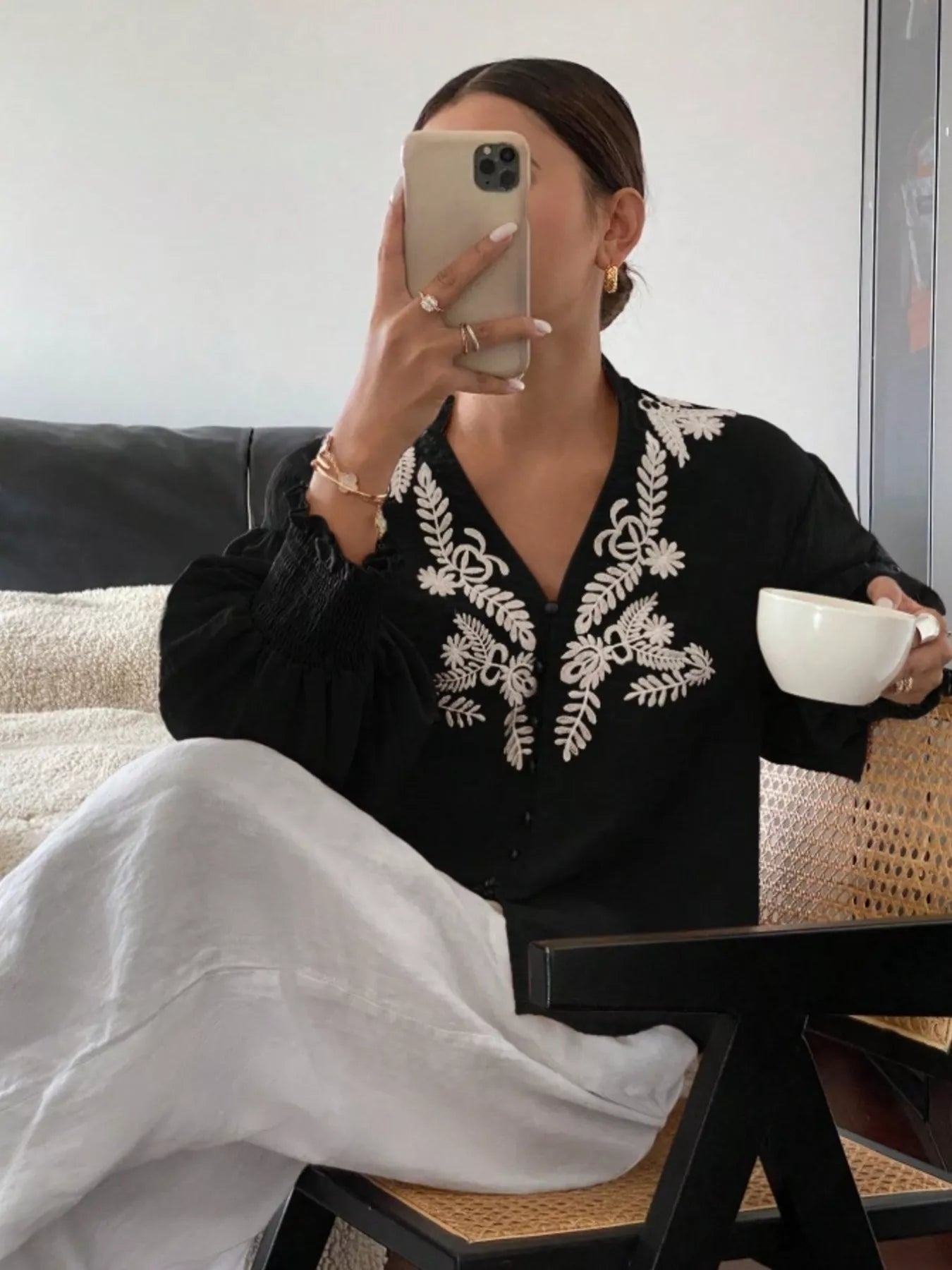 Person wearing a black embroidered top sitting on a couch holding a white cup.