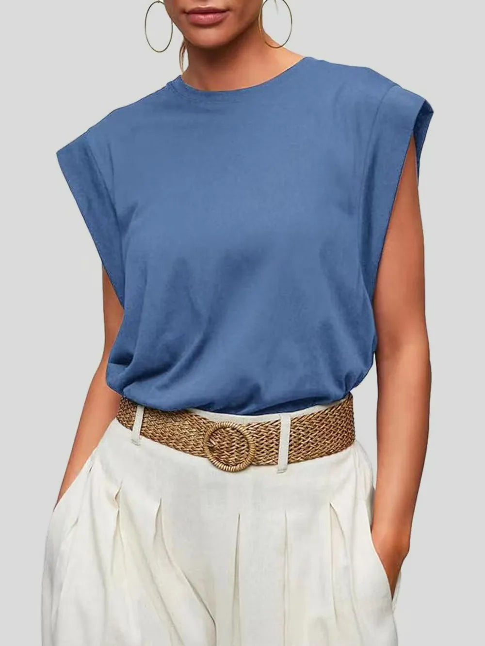 Person wearing a blue sleeveless top and white pants with a woven belt on a plain background