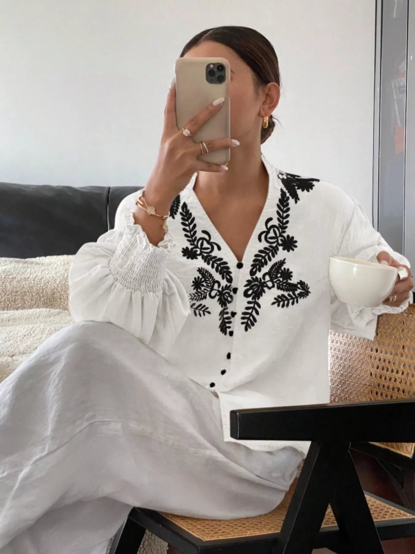 Woman in a white dress with black embroidery holding a cup, sitting on a couch.