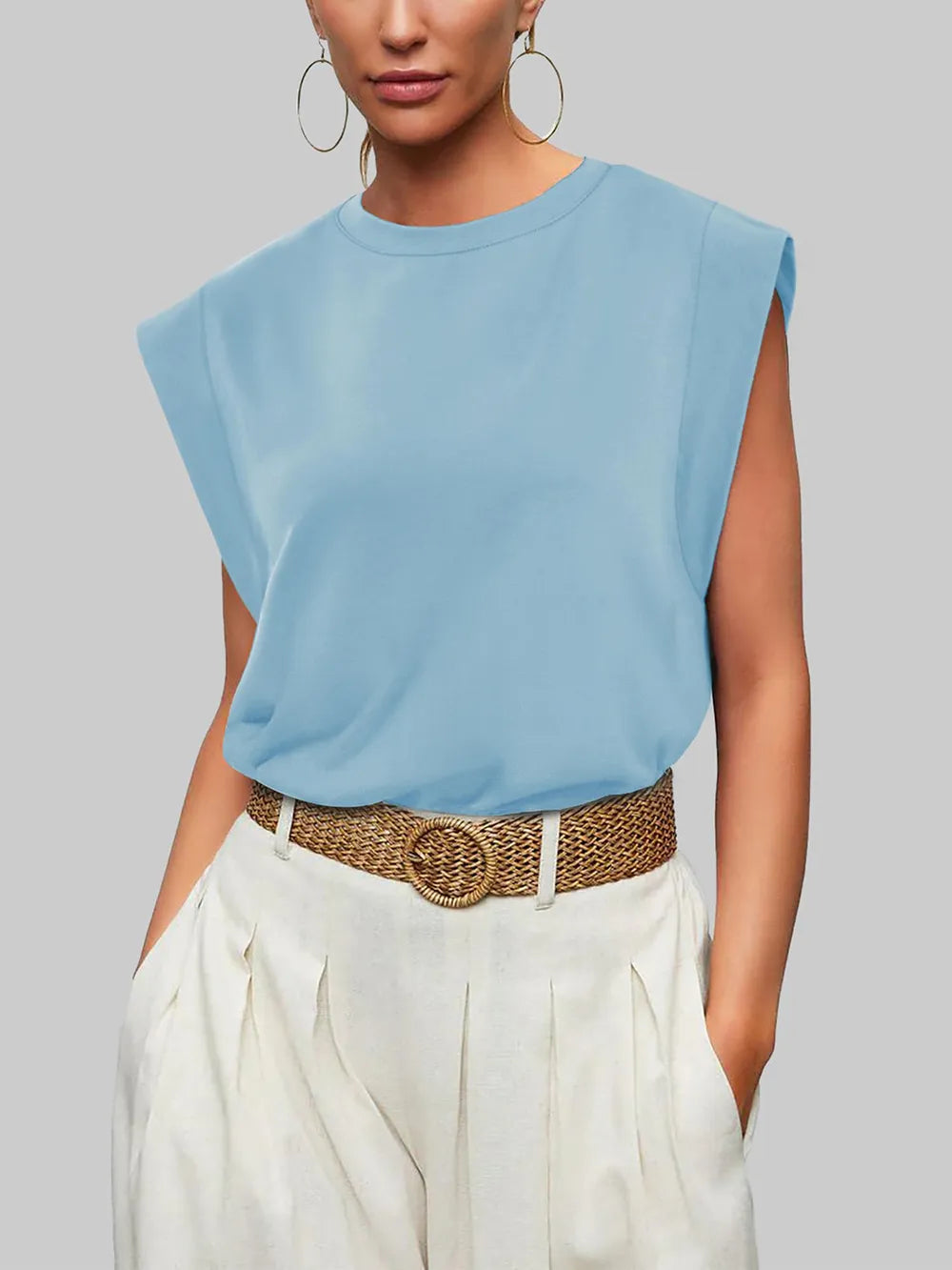 Woman wearing a light blue sleeveless top and white pants with a brown belt against a gray background