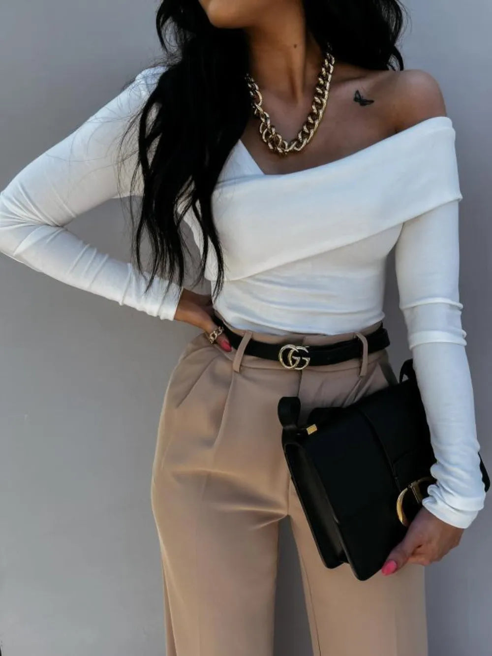 Woman wearing a white off-shoulder top and beige pants, holding a black handbag against a gray background.
