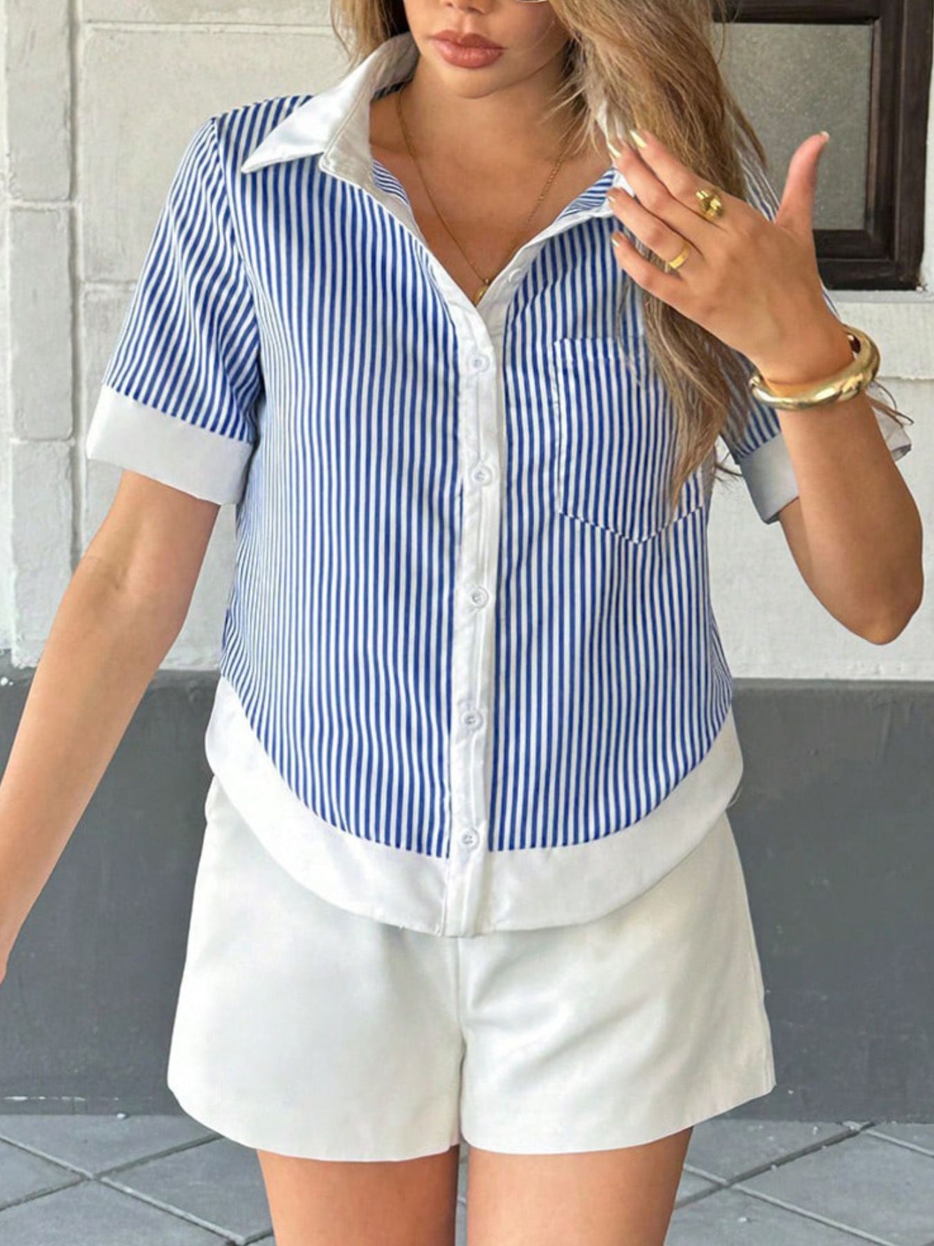Woman wearing a blue and white striped shirt and white shorts, standing against a gray wall.