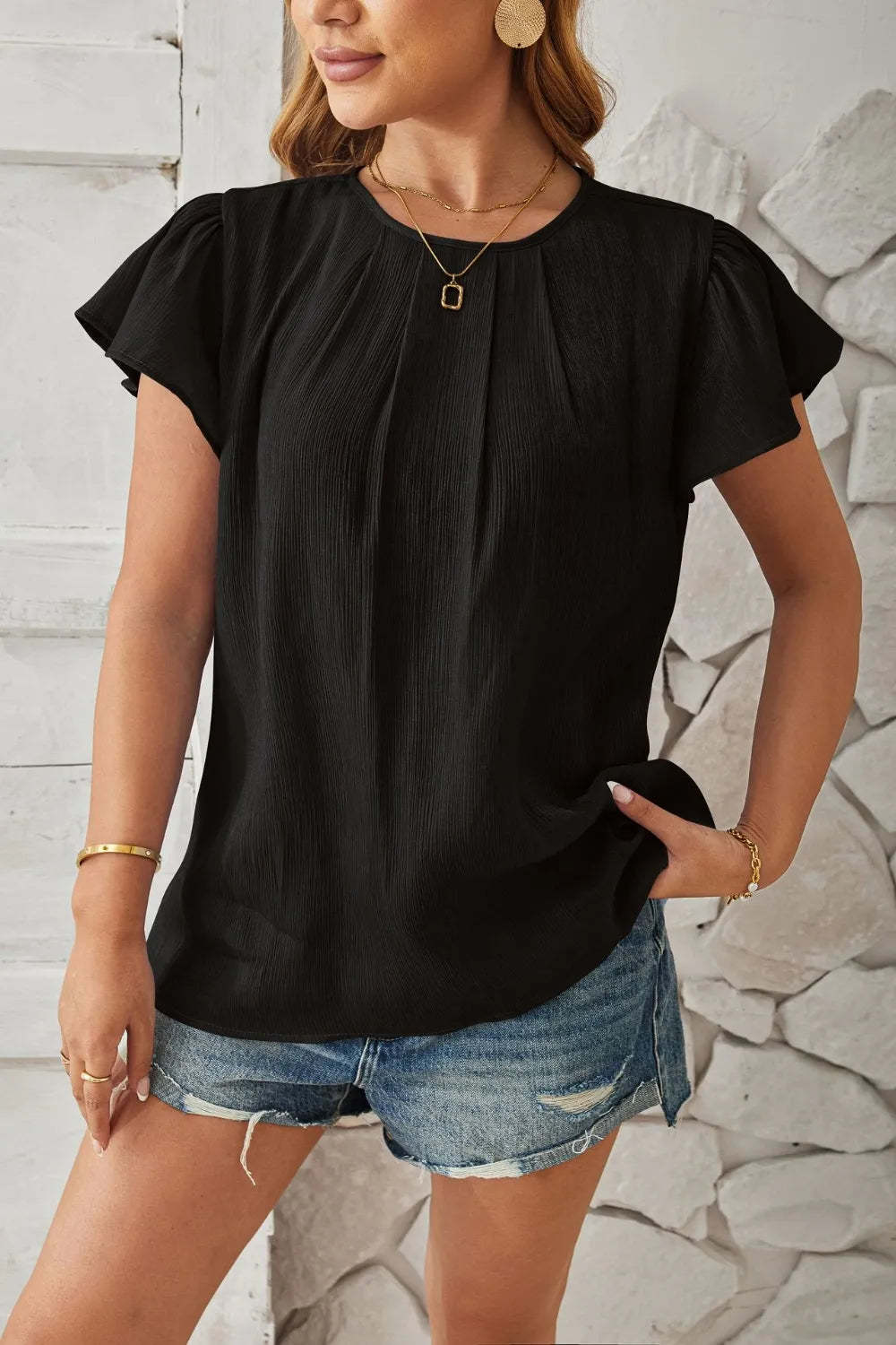 Woman wearing a black top and denim shorts against a stone wall.